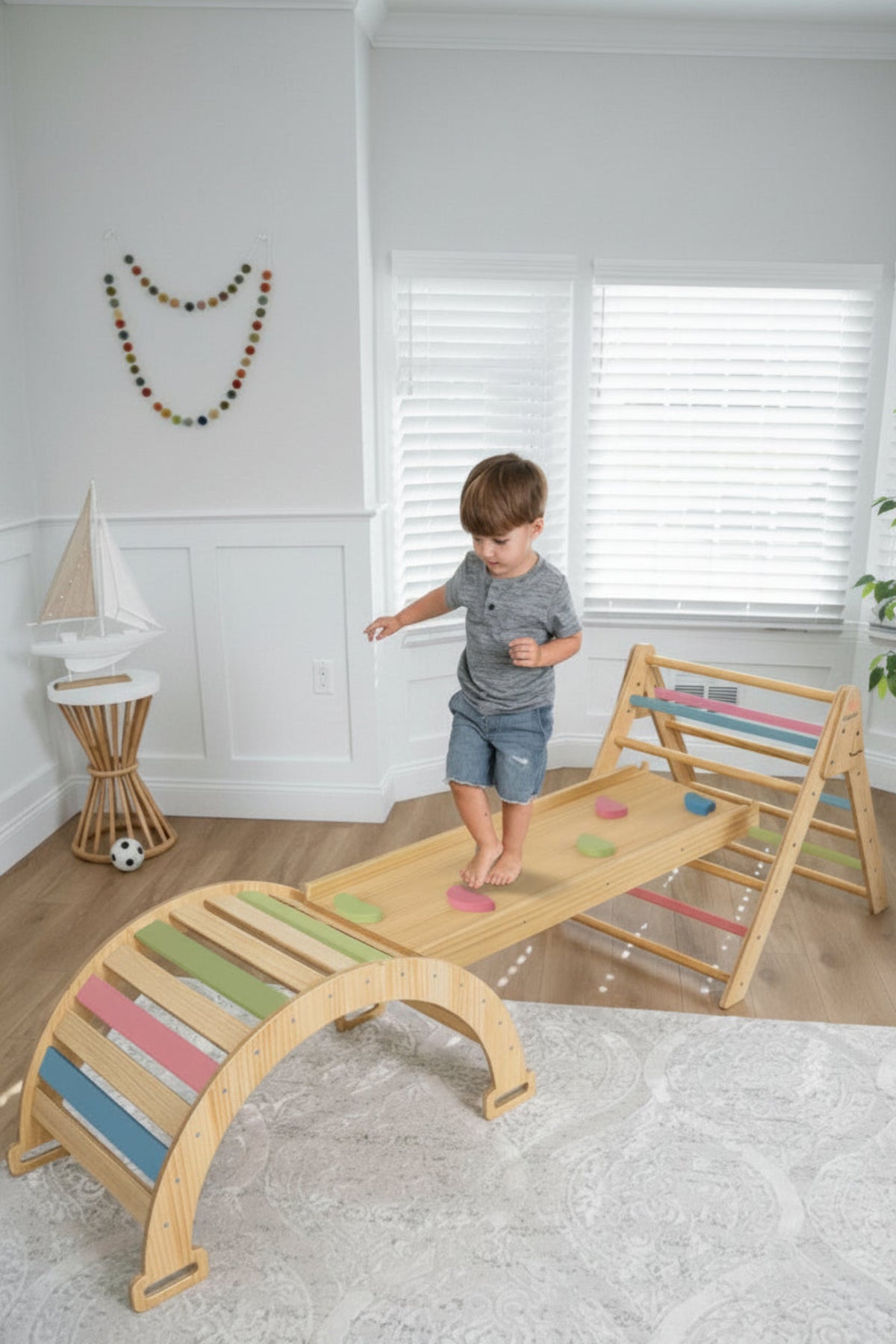 A young child balances on a wooden Pikler triangle playset featuring colorful climbing elements. The playset is set in a bright, airy room with natural light.