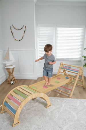 A young child balances on a wooden Pikler triangle playset featuring colorful climbing elements. The playset is set in a bright, airy room with natural light.