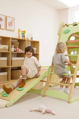 Two children play on a wooden jungle gym with a slide and climbing wall in a bright, minimalist playroom. The set features a soft green finish, promoting active indoor play.