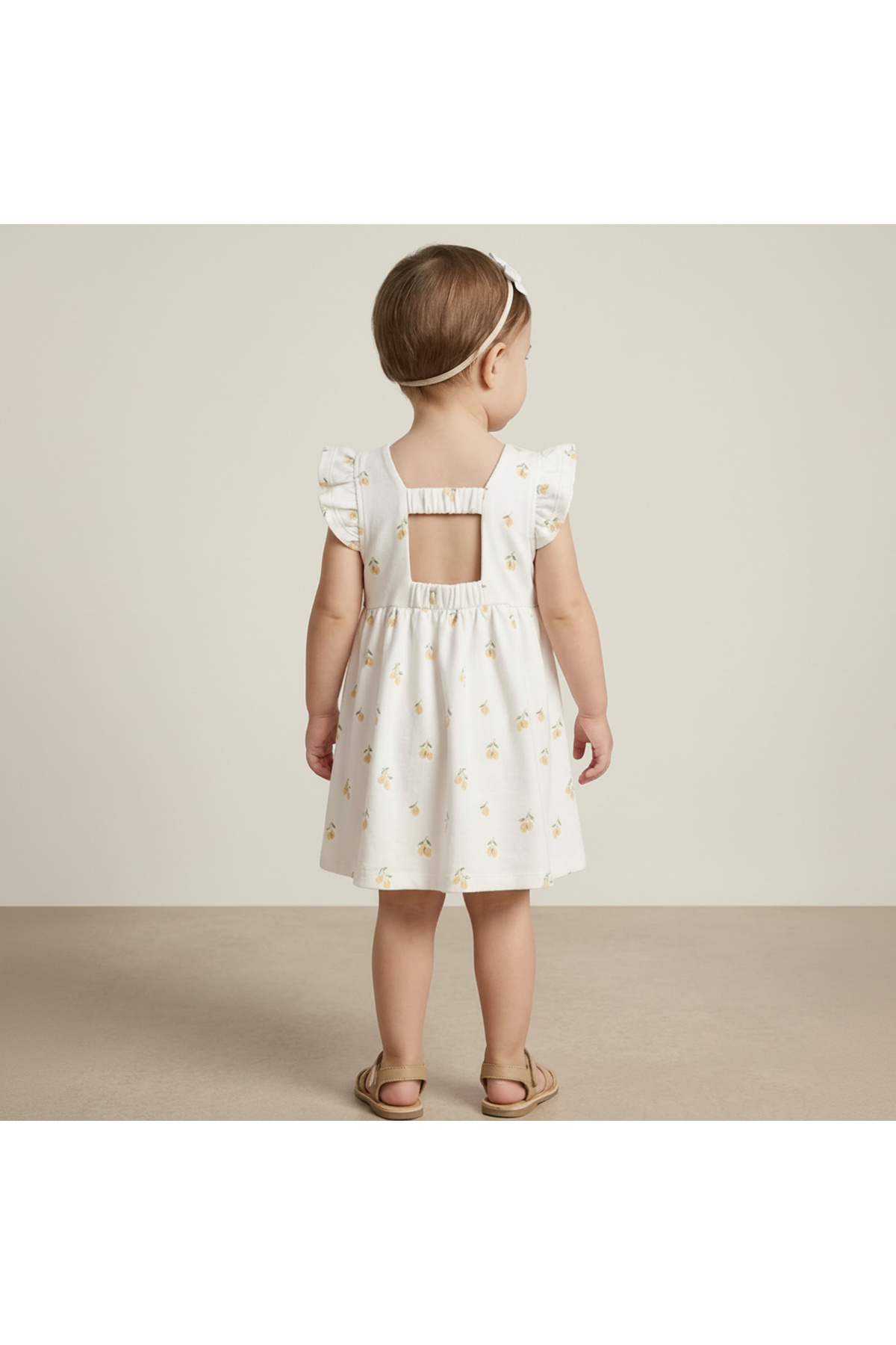 A young child stands with their back to the camera, wearing a white dress with a cutout back and lemon print.