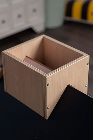 A wooden storage box with a light finish sits on a black tabletop, featuring a slotted design and smooth edges. The background shows a bright, organized playroom setting.