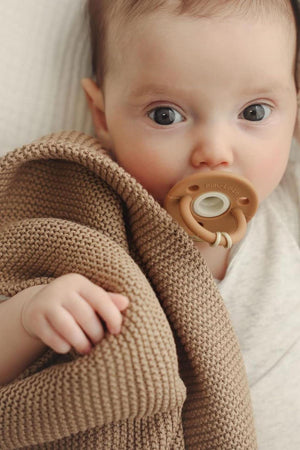 A baby wrapped in a brown knit blanket with a beige pacifier, lying on a light-colored surface.