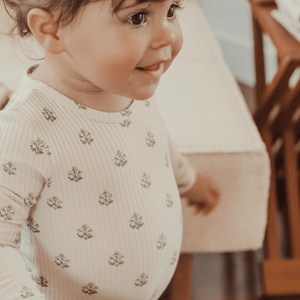 A smiling toddler wearing a light-colored ribbed onesie with a delicate floral pattern stands beside a wooden chair. The soft fabric and charming design create a cozy, playful look.
