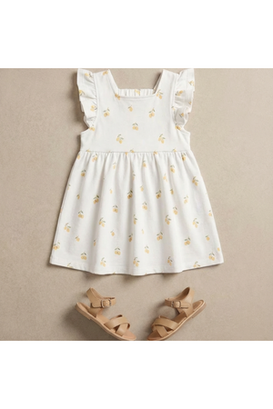 A white dress with a lemon print and ruffled sleeves is displayed on a neutral background, paired with light brown sandals.