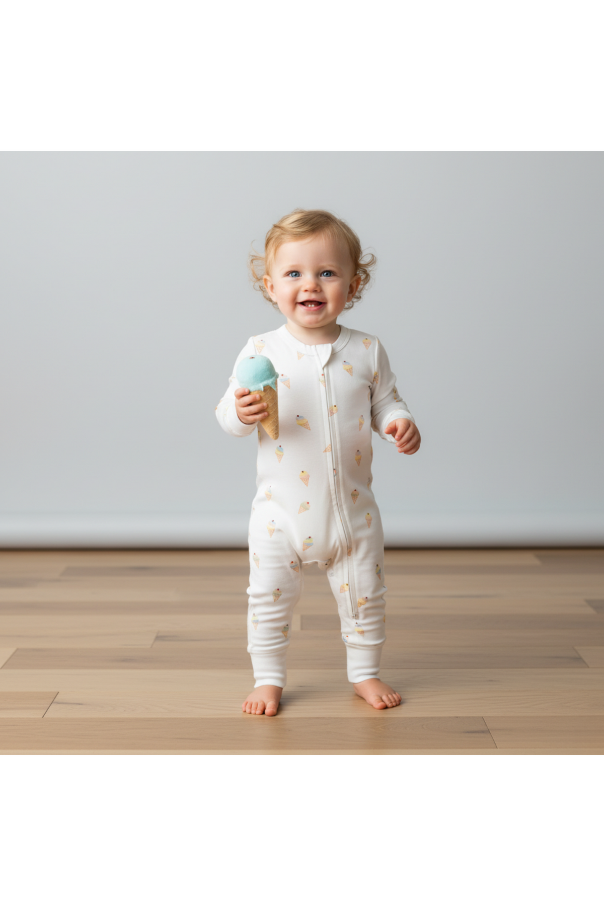 A smiling toddler wearing an organic cotton romper with an ice cream cone print stands on a wooden floor, holding a toy ice cream cone.