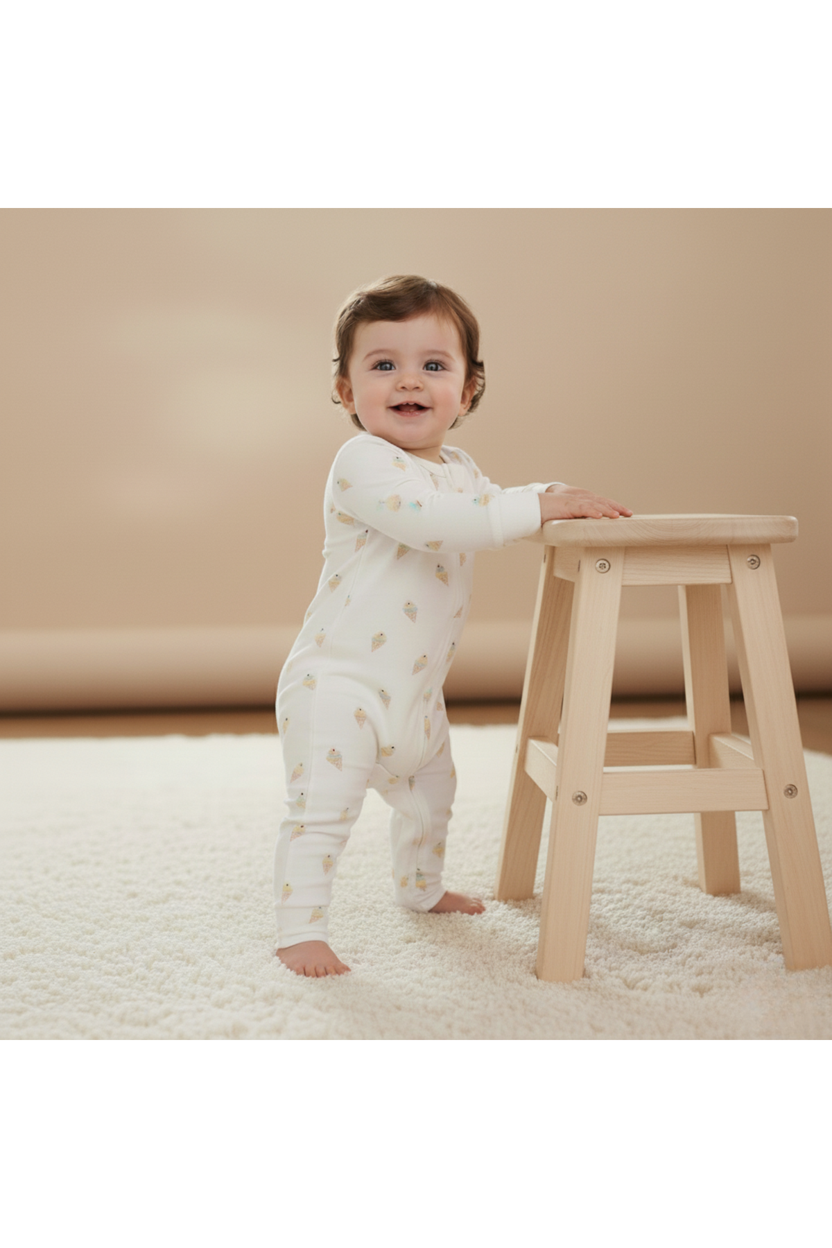 A smiling baby stands beside a wooden stool, wearing a soft, organic cotton romper adorned with ice cream cone prints.