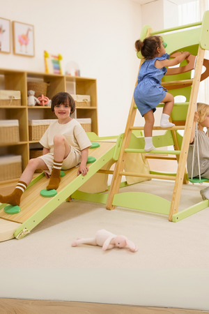 Two children play on the Bush 6 in 1 Wooden Jungle Gym Climber, featuring a green slide and climbing wall, in a bright, spacious playroom with wooden storage units.