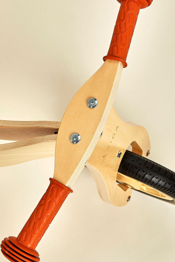 Close-up view of a wooden balance bike featuring a light-colored wooden frame and bright orange rubber grips.