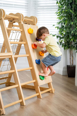 A toddler climbs a wooden jungle gym with colorful holds indoors, near a leafy plant.
