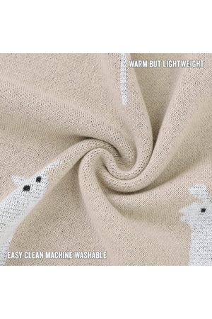 Beige knit blanket with white llama pattern, twisted to display texture. Text reads Warm but lightweight, easy clean machine washable.