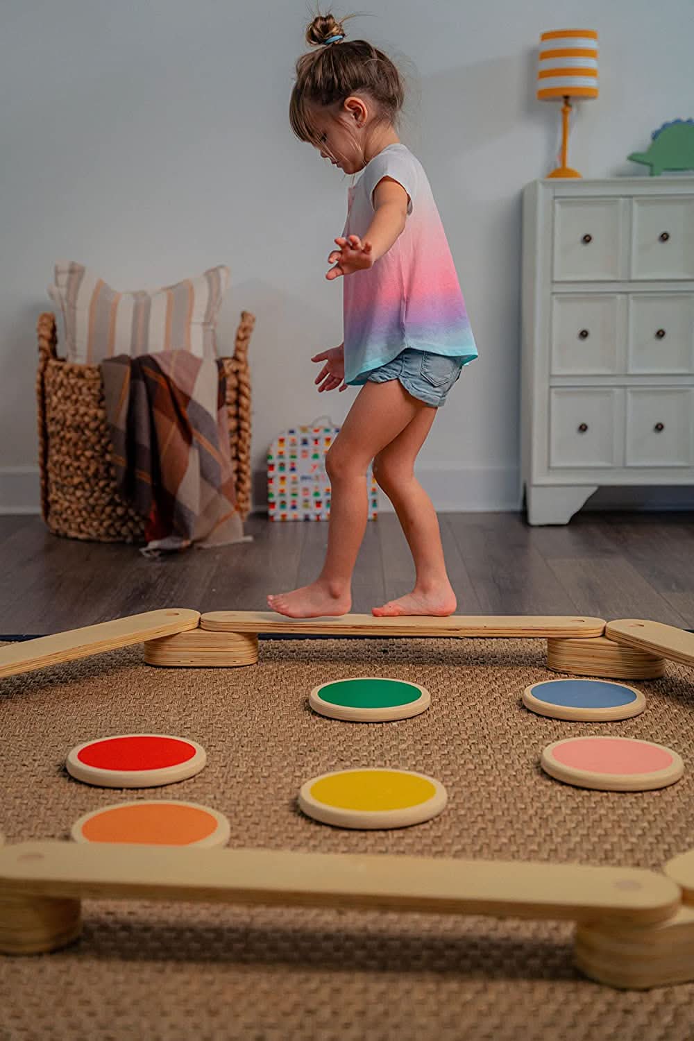 A young girl balances on a wooden beam above colorful stepping stones in a bright, inviting playroom.