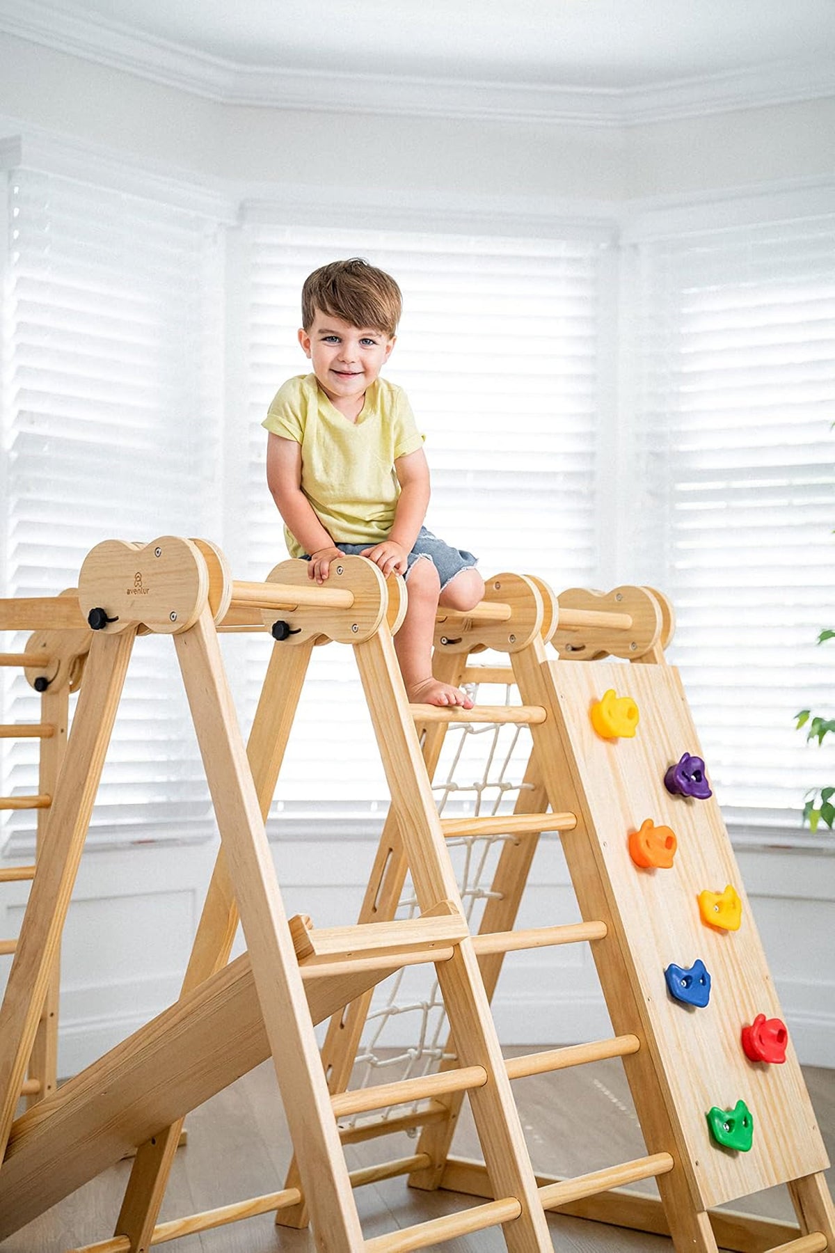 A child sits on a wooden jungle gym with climbing features, in a bright room with white blinds.