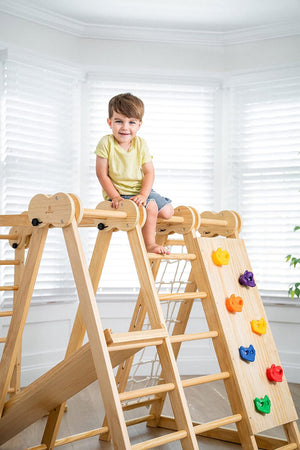 A child sits on a wooden jungle gym with climbing features, in a bright room with white blinds.