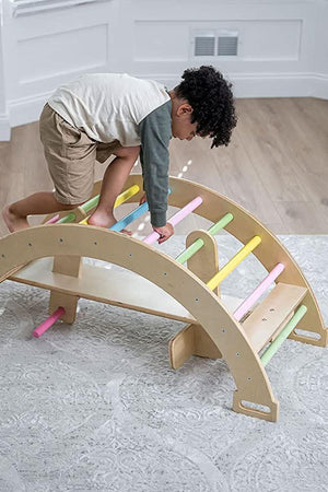 A child with curly hair climbs on a wooden climbing arch with colorful, non-toxic bars, set on a light gray patterned rug in a bright, airy room.