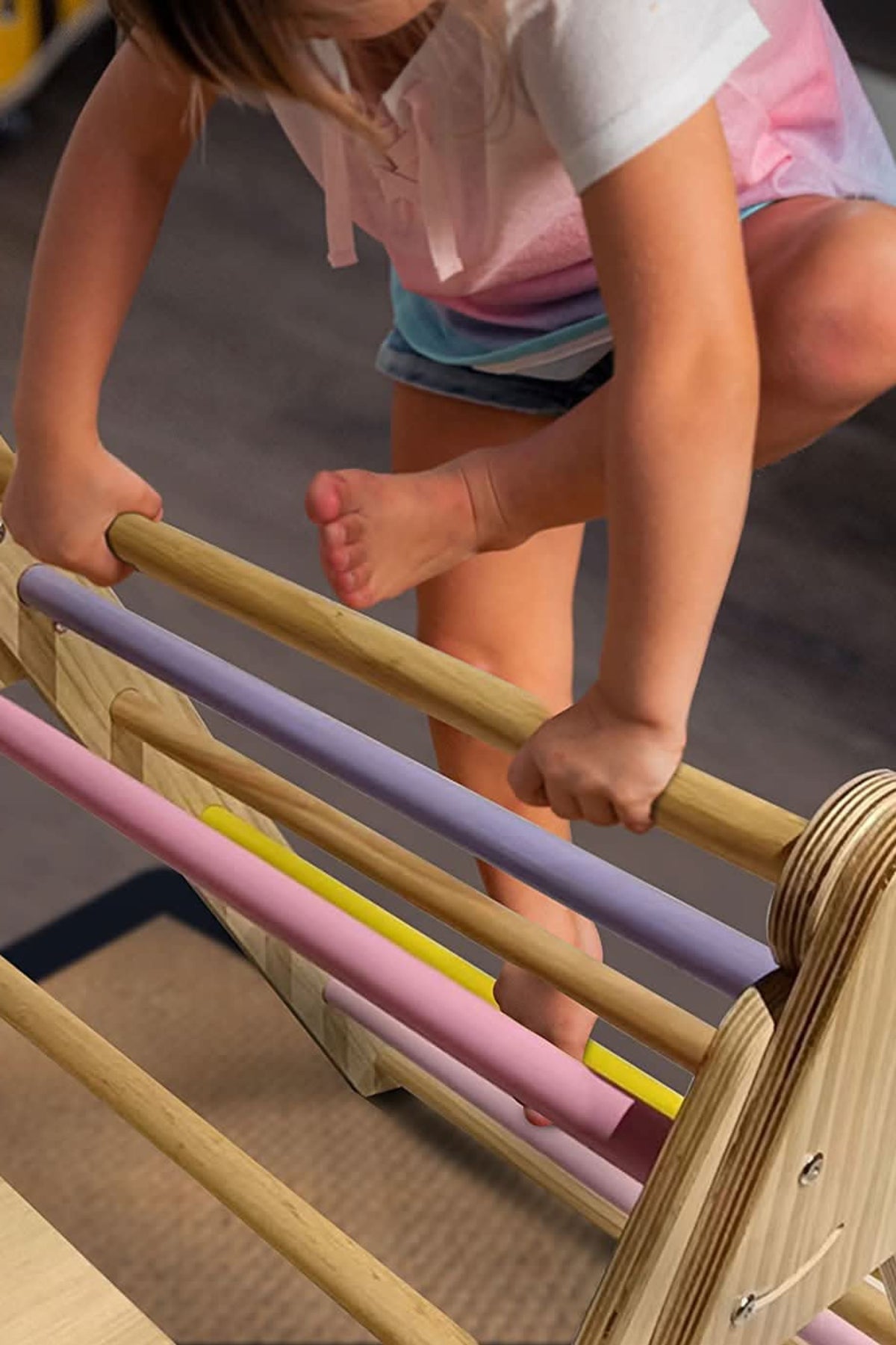 A child climbs on the Hazel Pikler Triangle, showcasing its wooden structure and colorful, padded rungs in pastel shades. The setting is indoors, emphasizing safe and active play.