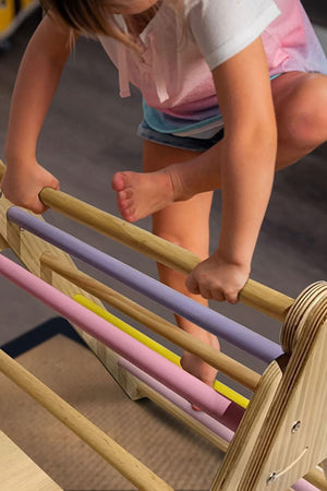 A child climbs on the Hazel Pikler Triangle, showcasing its wooden structure and colorful, padded rungs in pastel shades. The setting is indoors, emphasizing safe and active play.