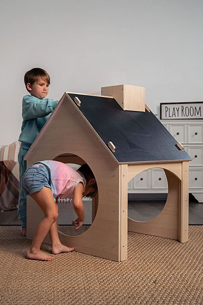 A boy and girl play with an evergreen wooden childrens playhouse featuring a chalkboard roof in a cozy playroom. The house is made from sustainable materials, promoting imaginative play.