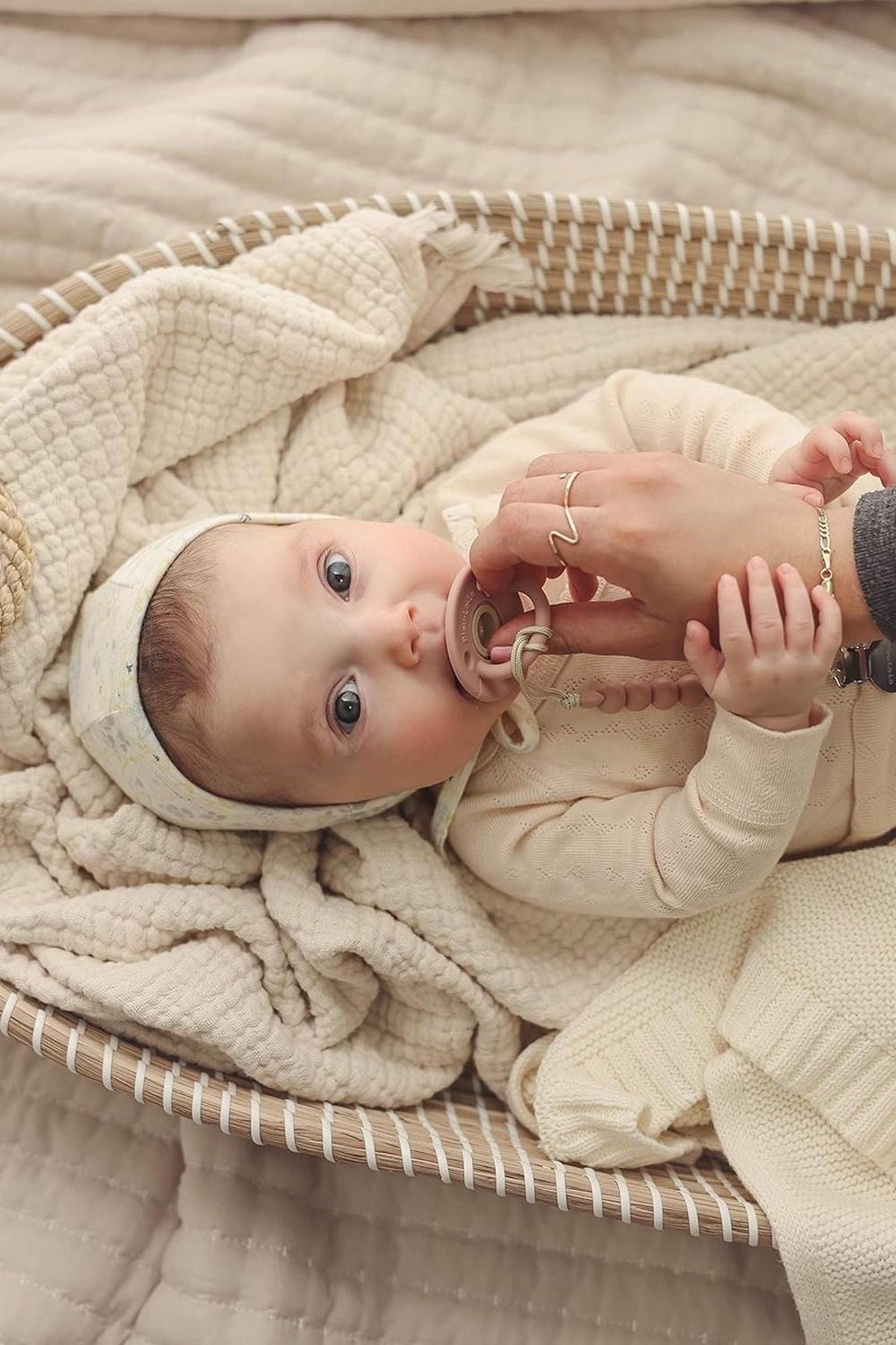 A baby lies in a woven basket with a pacifier, wrapped in a soft, cream-colored knit blanket.