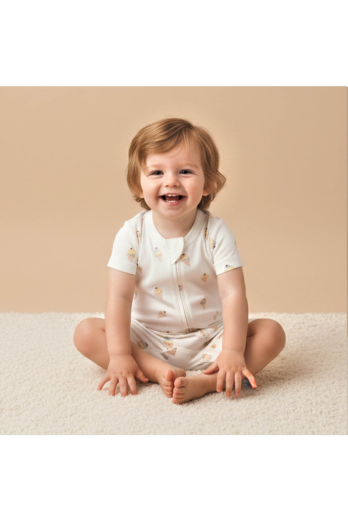 A smiling child sits on a soft, light-colored carpet, wearing an organic cotton short zip romper adorned with a playful ice cream print, perfect for sunny days and adventures.