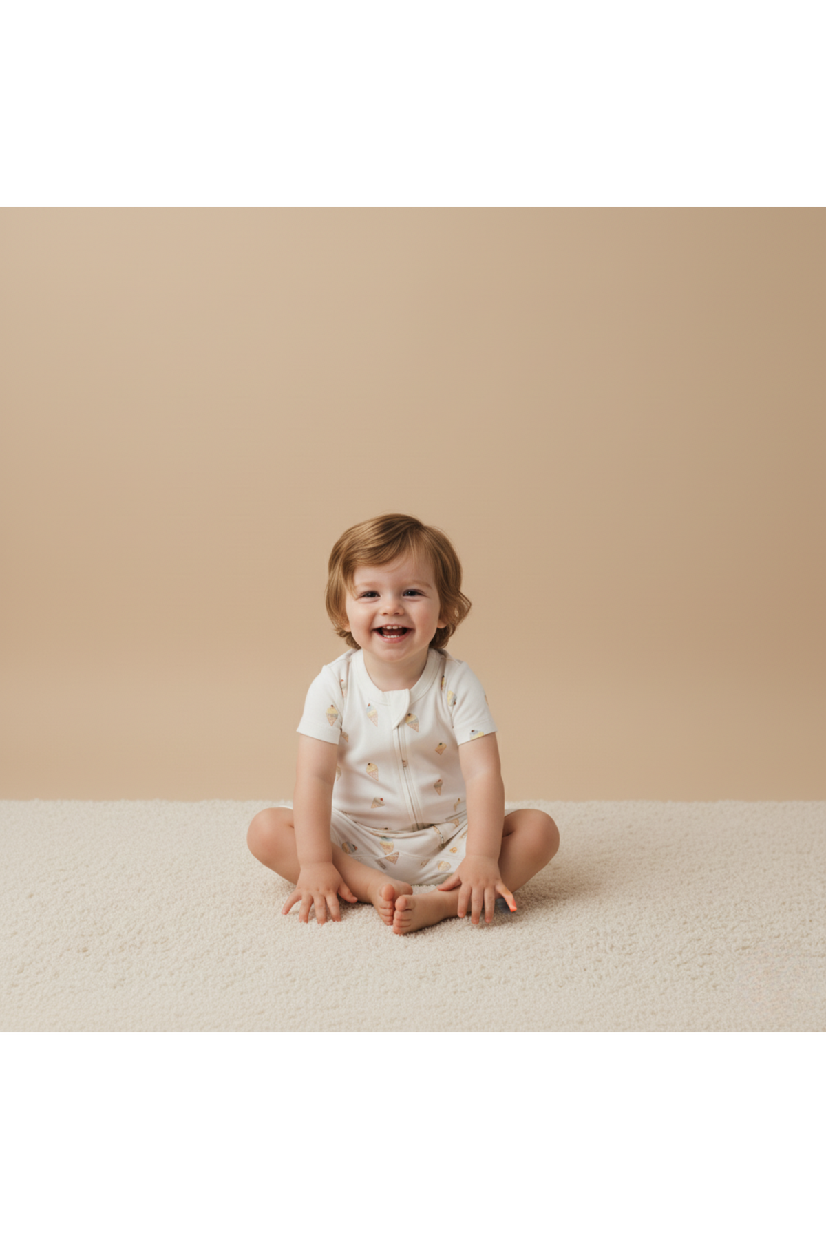 A smiling toddler sits on a soft, light-colored rug, wearing a white short-sleeve romper adorned with subtle patterns. The backdrop is a warm beige, enhancing the playful atmosphere.