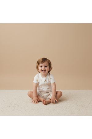 A smiling toddler sits on a soft, light-colored rug, wearing a white short-sleeve romper adorned with subtle patterns. The backdrop is a warm beige, enhancing the playful atmosphere.
