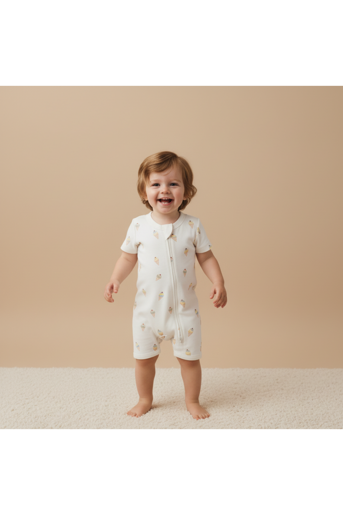 A smiling toddler stands on a soft, textured surface, wearing a short-sleeved, cream-colored romper with subtle ice cream cone patterns. The outfit features a convenient front zip for easy dressing.