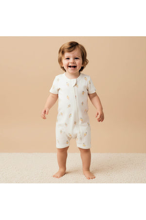 A smiling toddler stands on a soft, cream-colored rug, wearing an organic cotton short zip romper featuring a playful ice cream print. The background is a warm, neutral tone.