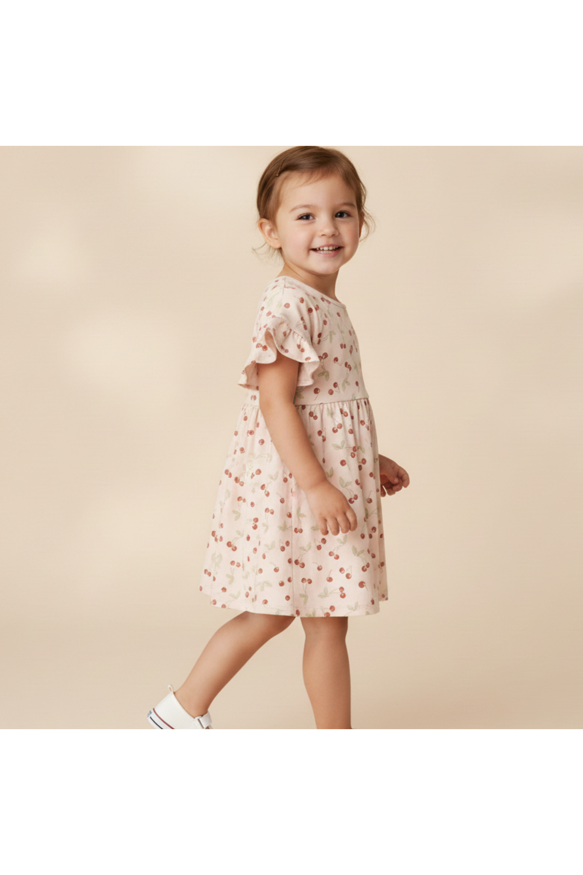 A young girl smiles while wearing a light pink organic cotton dress with a cherry print and dolman sleeves. The dress features a relaxed fit, perfect for playful outings.