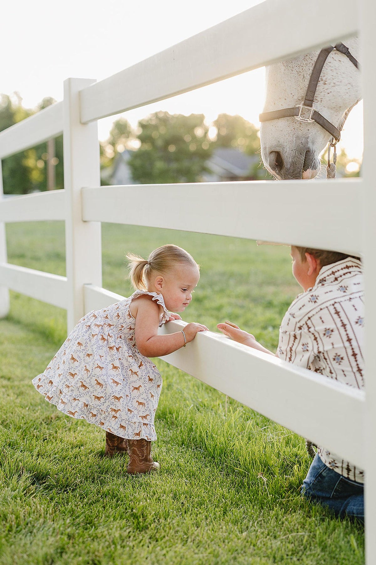 A young girl in a pony-printed smocked dress leans over a white fence, interacting with a horse, while a boy crouches beside her in a patterned shirt.