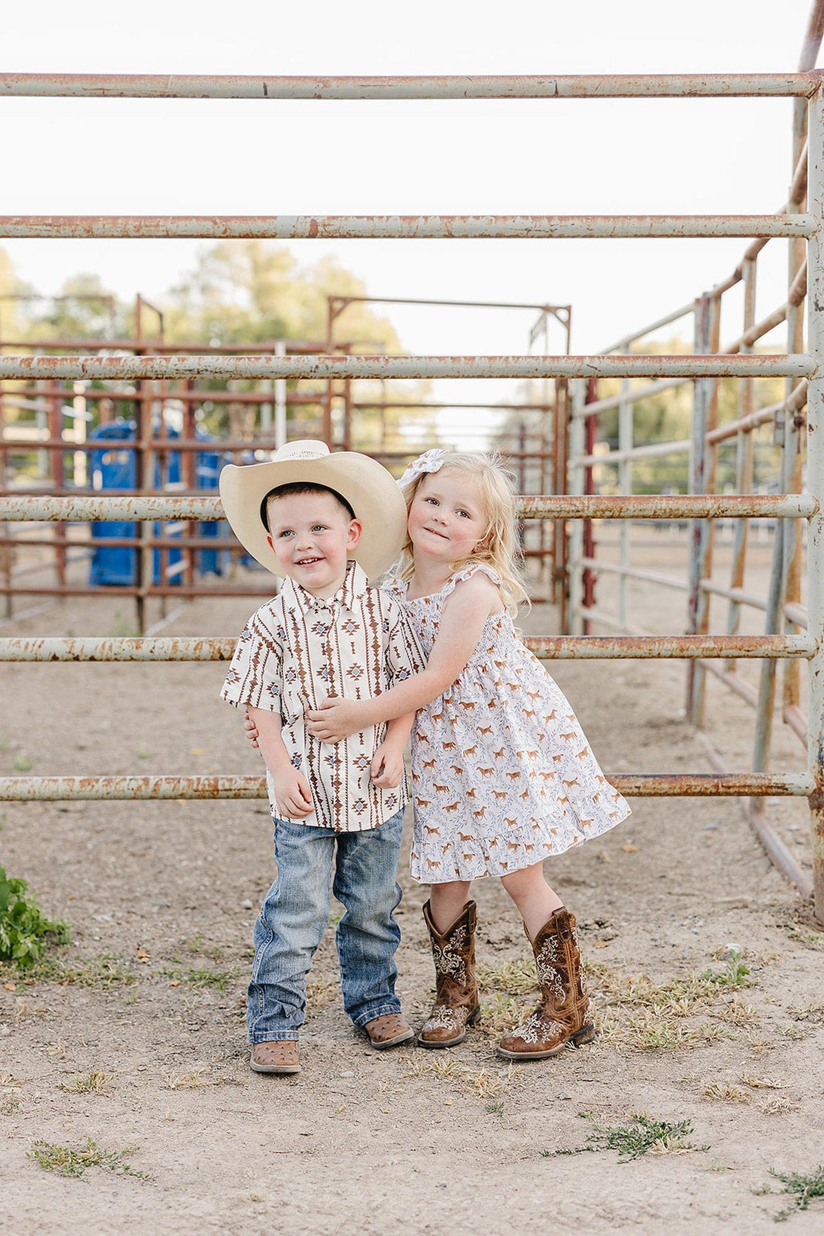 Two children stand together in a rustic outdoor setting, wearing cowboy attire. The girl is in a pony-printed dress and cowboy boots, while the boy sports a patterned shirt and.