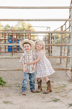 Two children stand together in a rustic outdoor setting, wearing cowboy attire. The girl is in a pony-printed dress and cowboy boots, while the boy sports a patterned shirt and.