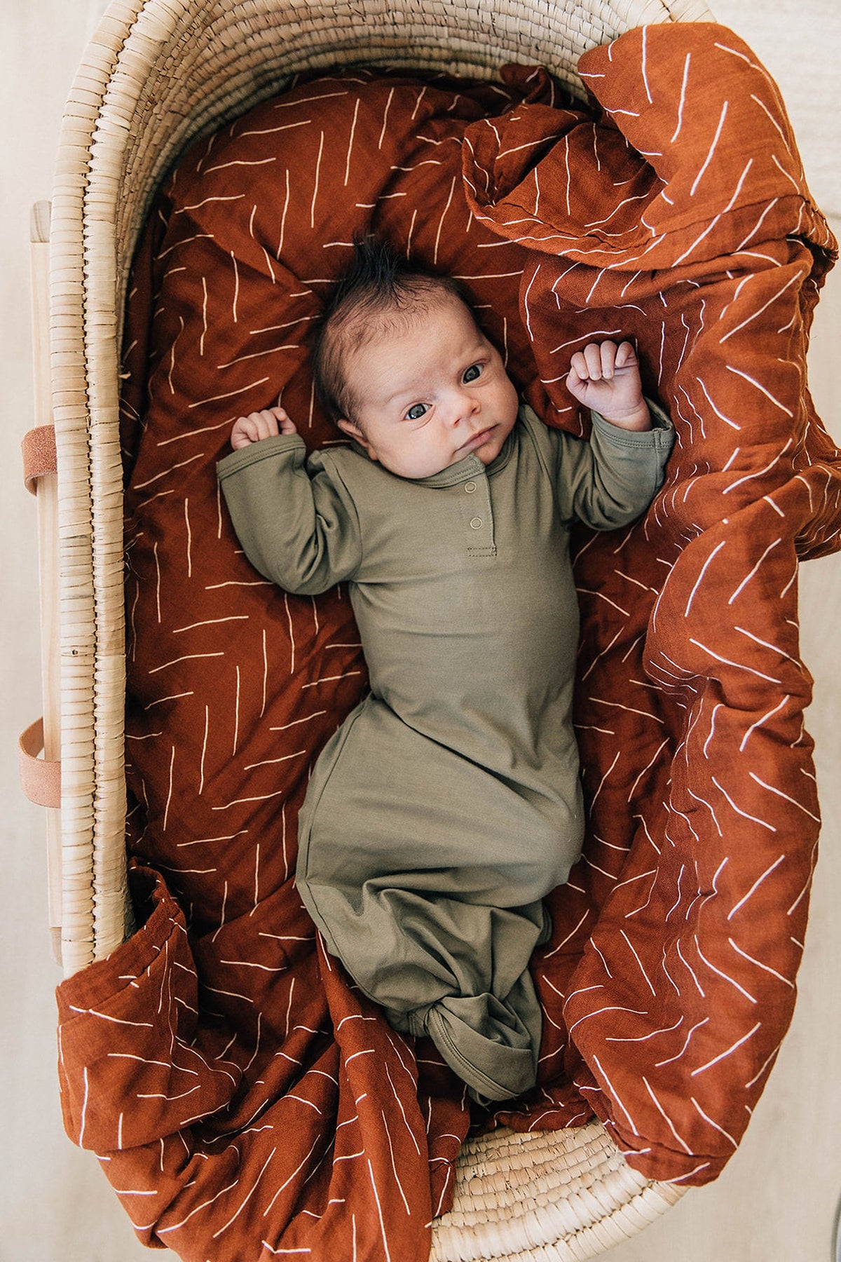 A baby in an olive green gown lies in a wicker basket with a rust-colored patterned blanket.