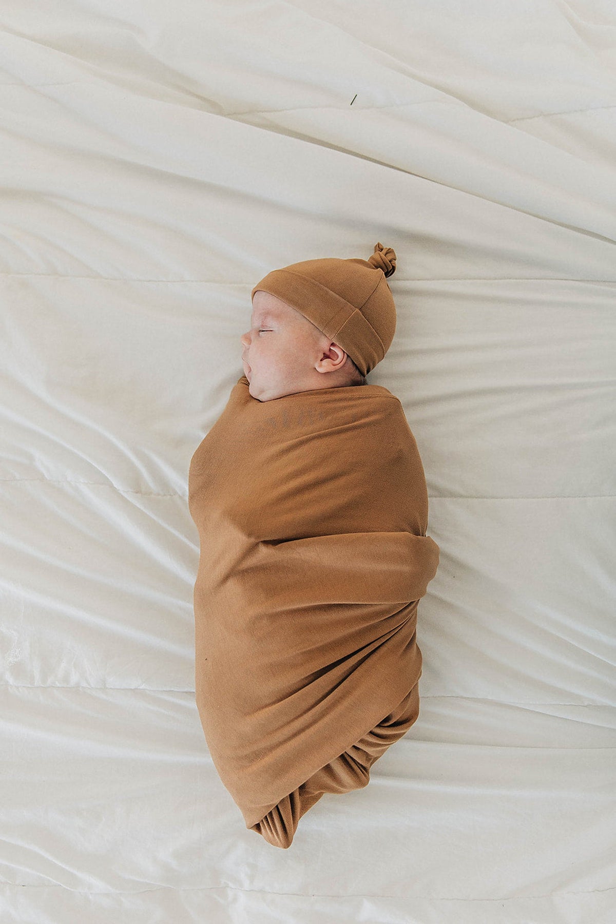 A newborn baby is swaddled in a soft, mustard-colored blanket with a matching knot hat, lying on a cream sheet.