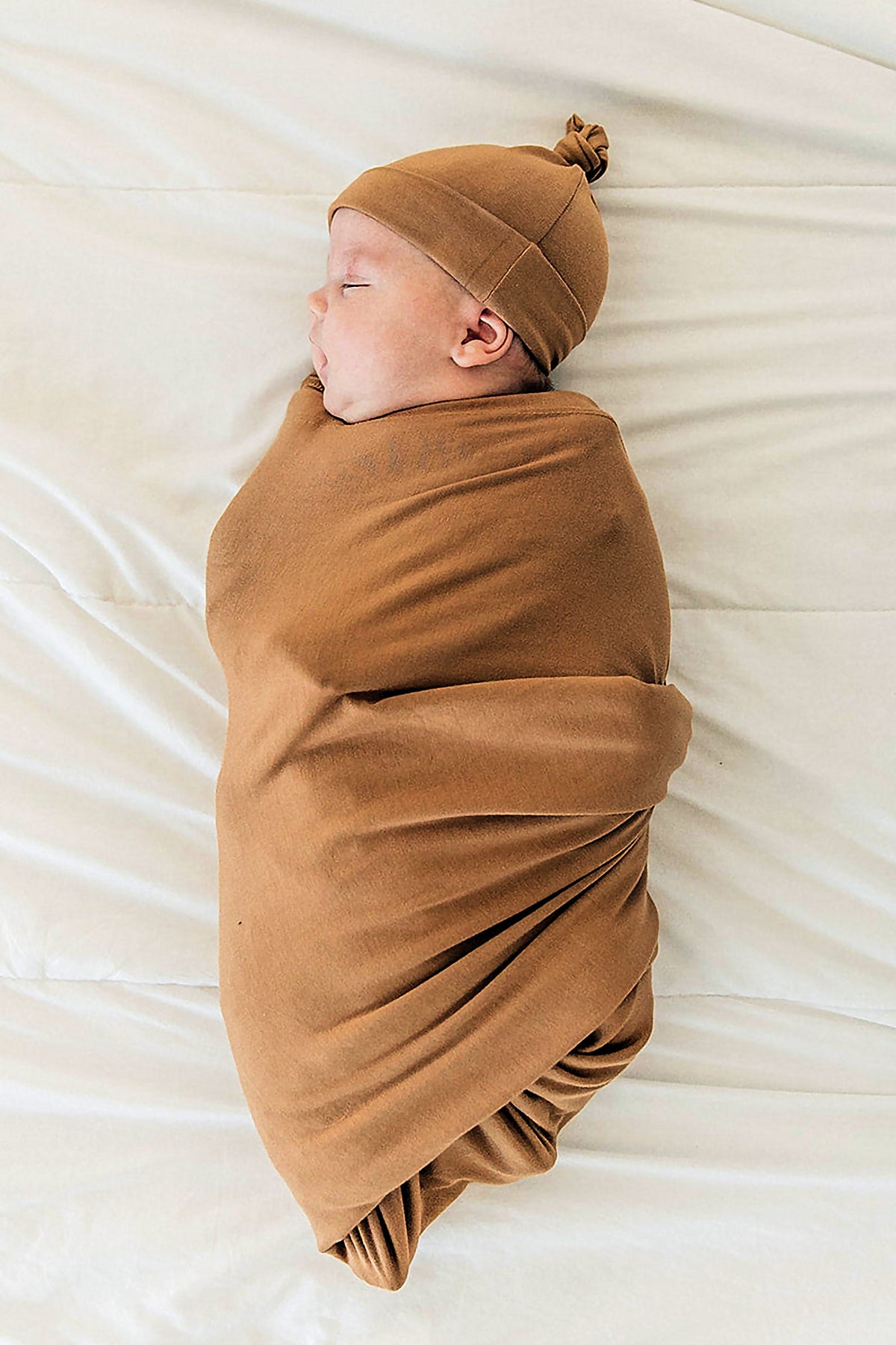 A baby is wrapped in a mustard-colored swaddle blanket, wearing a matching knotted hat, resting on a light surface.