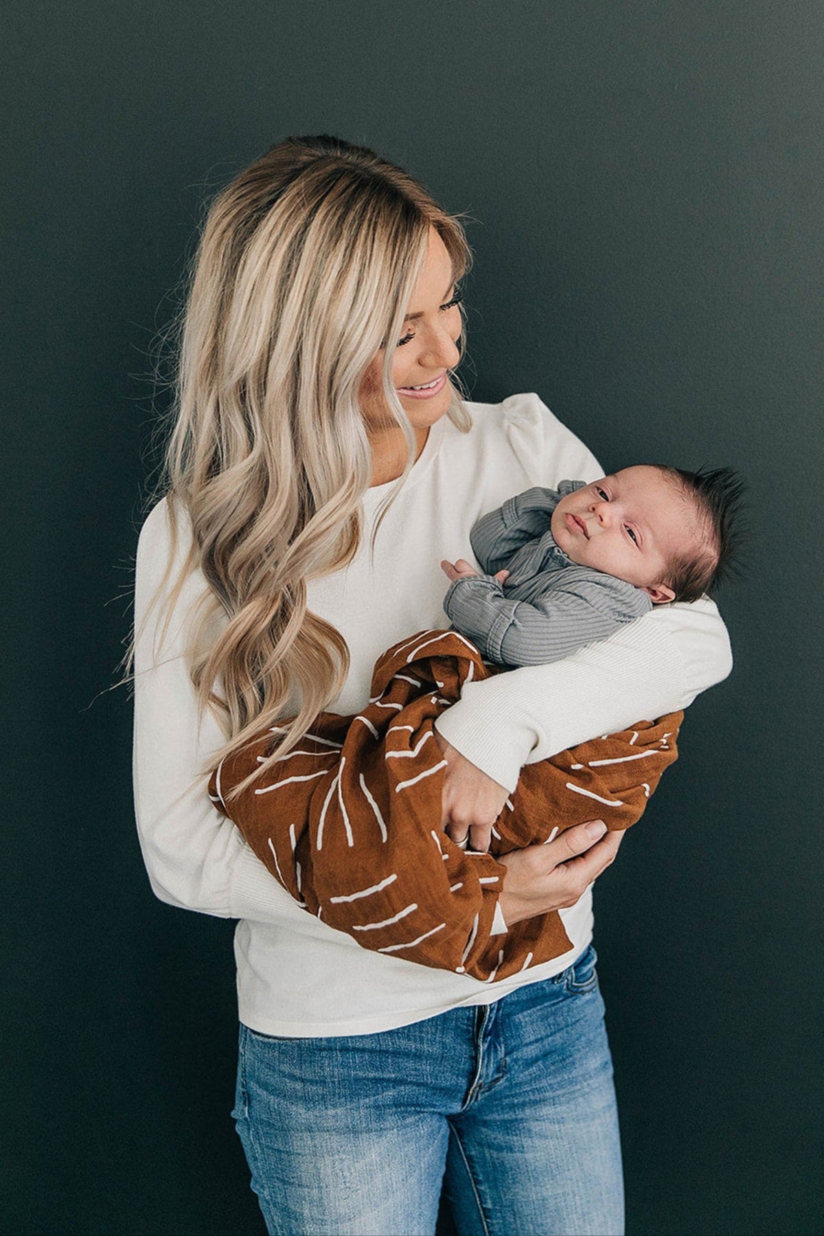 A woman in a white shirt holds a baby wrapped in a mustard-colored muslin swaddle with white patterns.