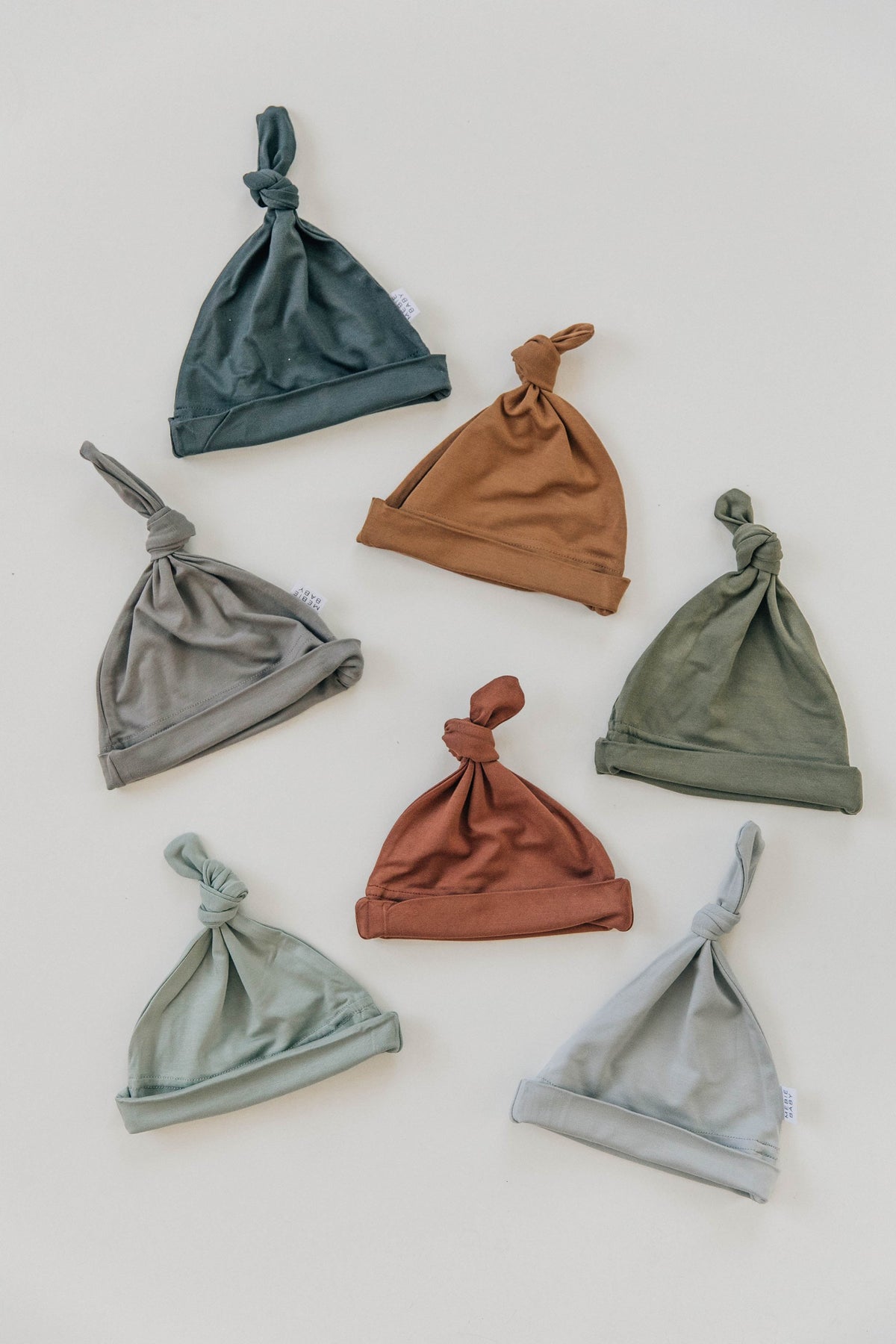 Seven newborn knot hats in earthy tones are arranged on a white background, showcasing soft bamboo fabric.