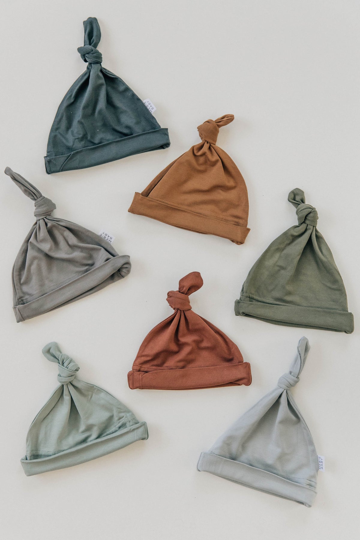 Seven newborn knot hats in various earthy tones, including rust, green, and blue, are arranged on a white background.