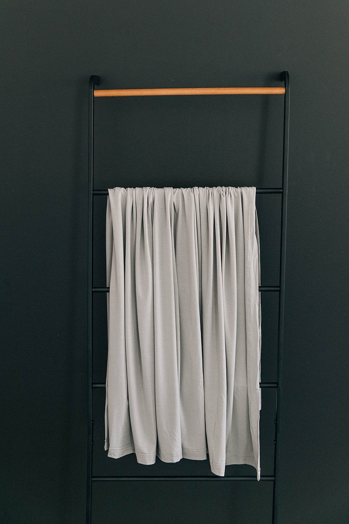 A light gray fabric swaddle hangs on a minimalist black metal rack against a dark wall.