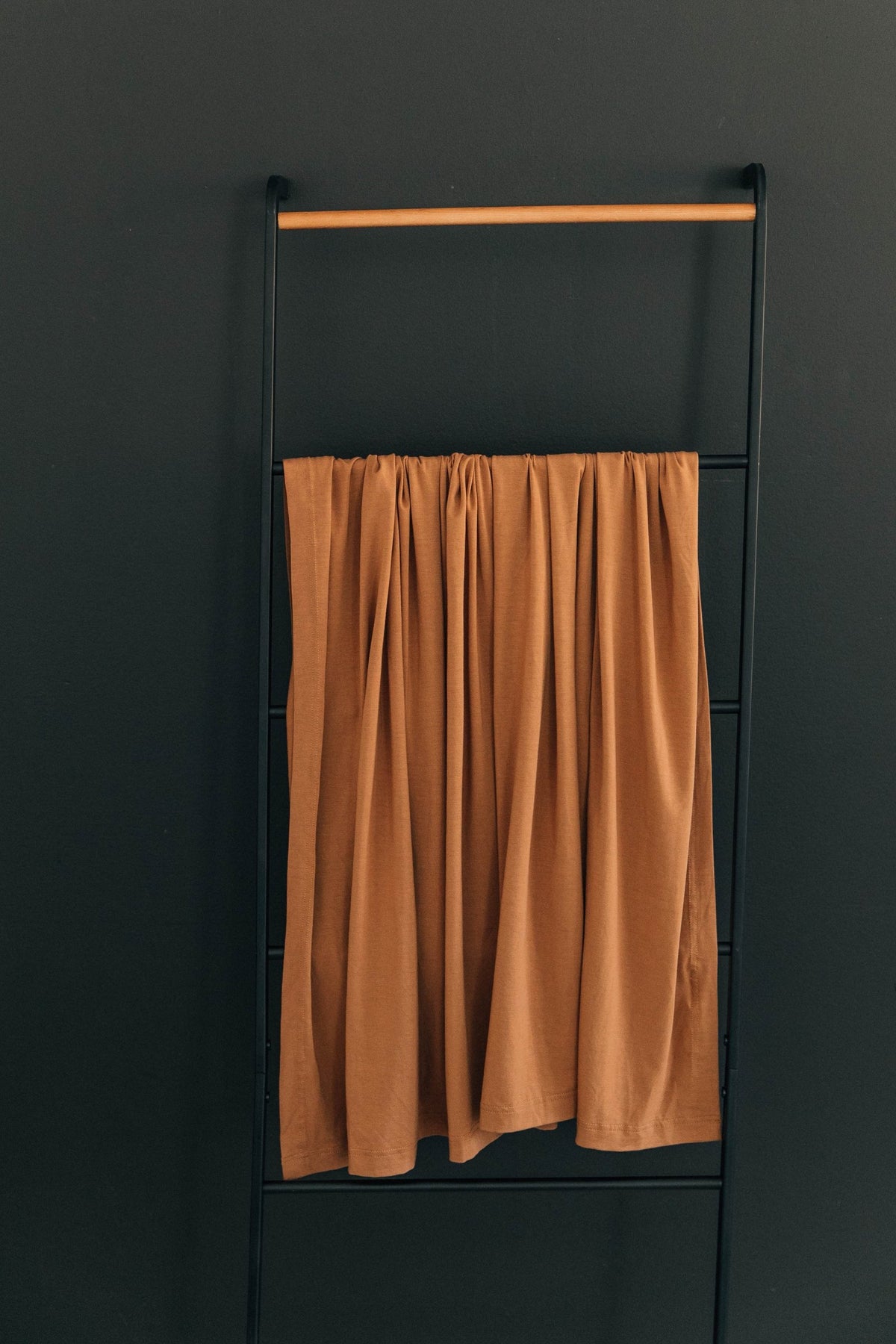 A mustard-colored swaddle blanket hangs on a black metal rack against a dark wall.
