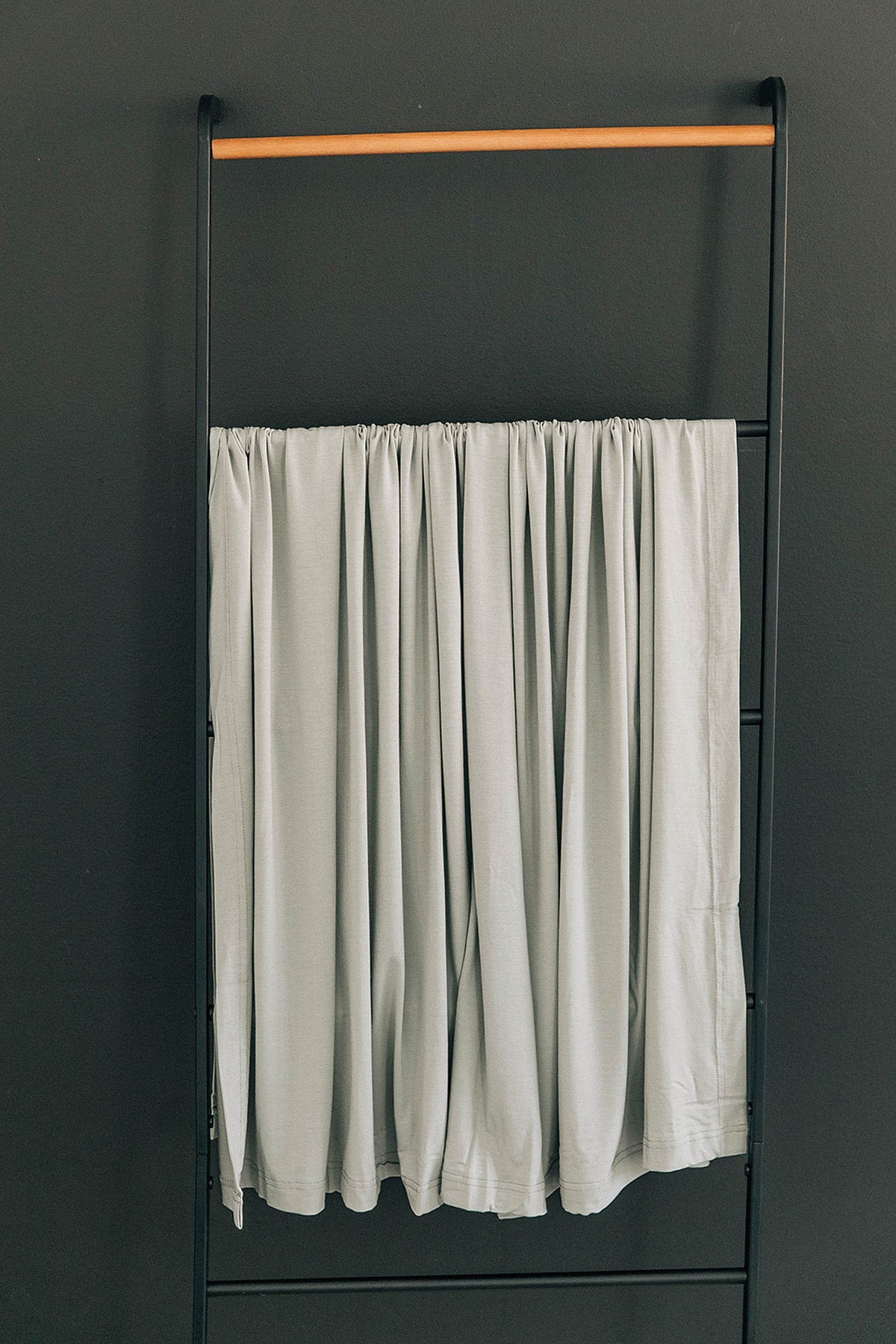 A sage-colored swaddle blanket hangs on a black metal rack with a wooden bar against a dark wall.