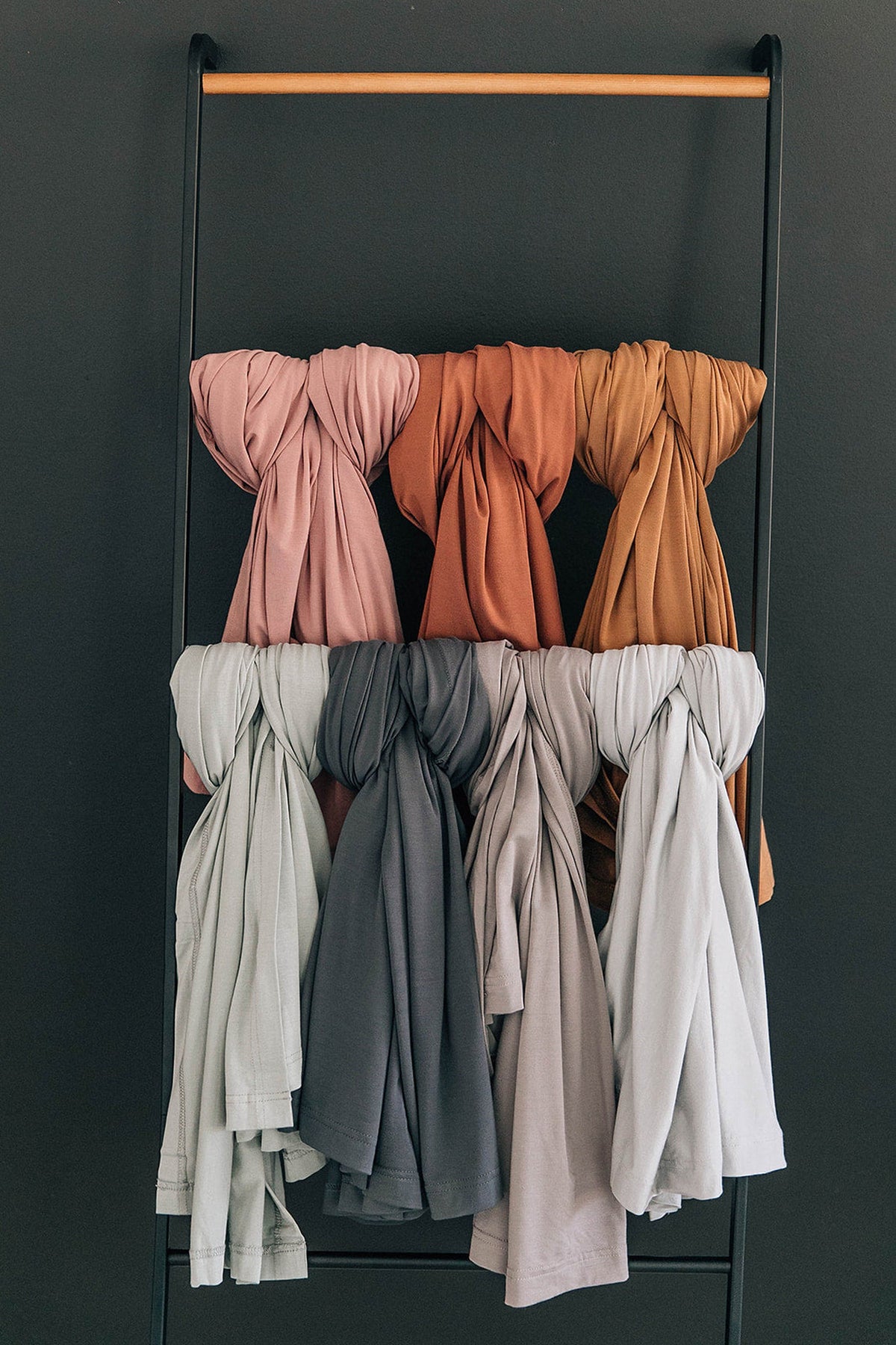 Six swaddle blankets in pink, orange, tan, sage, gray, and beige hang on a black metal rack.