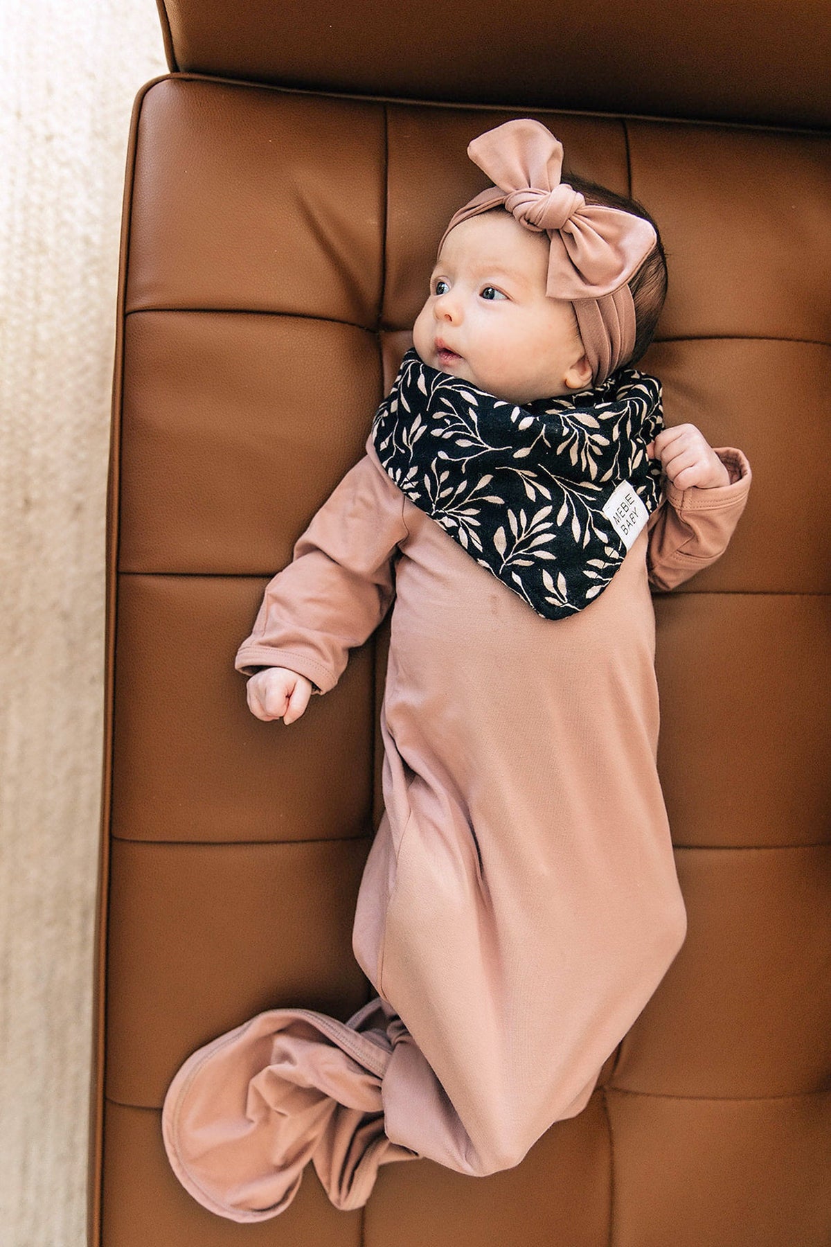 A baby wearing a dusty rose knot gown and matching headband lies on a brown leather couch.