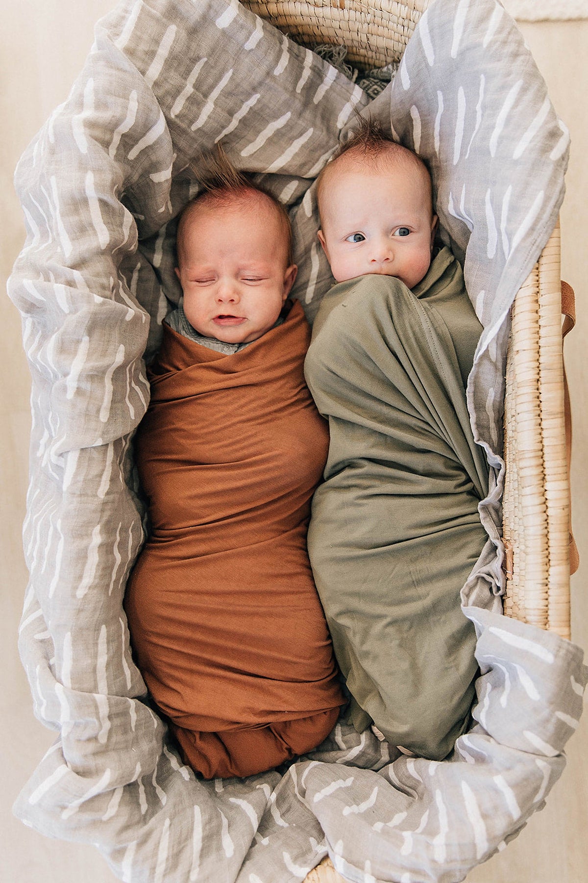 Two babies are swaddled in brown and olive stretch blankets, lying in a woven basket with a patterned lining.