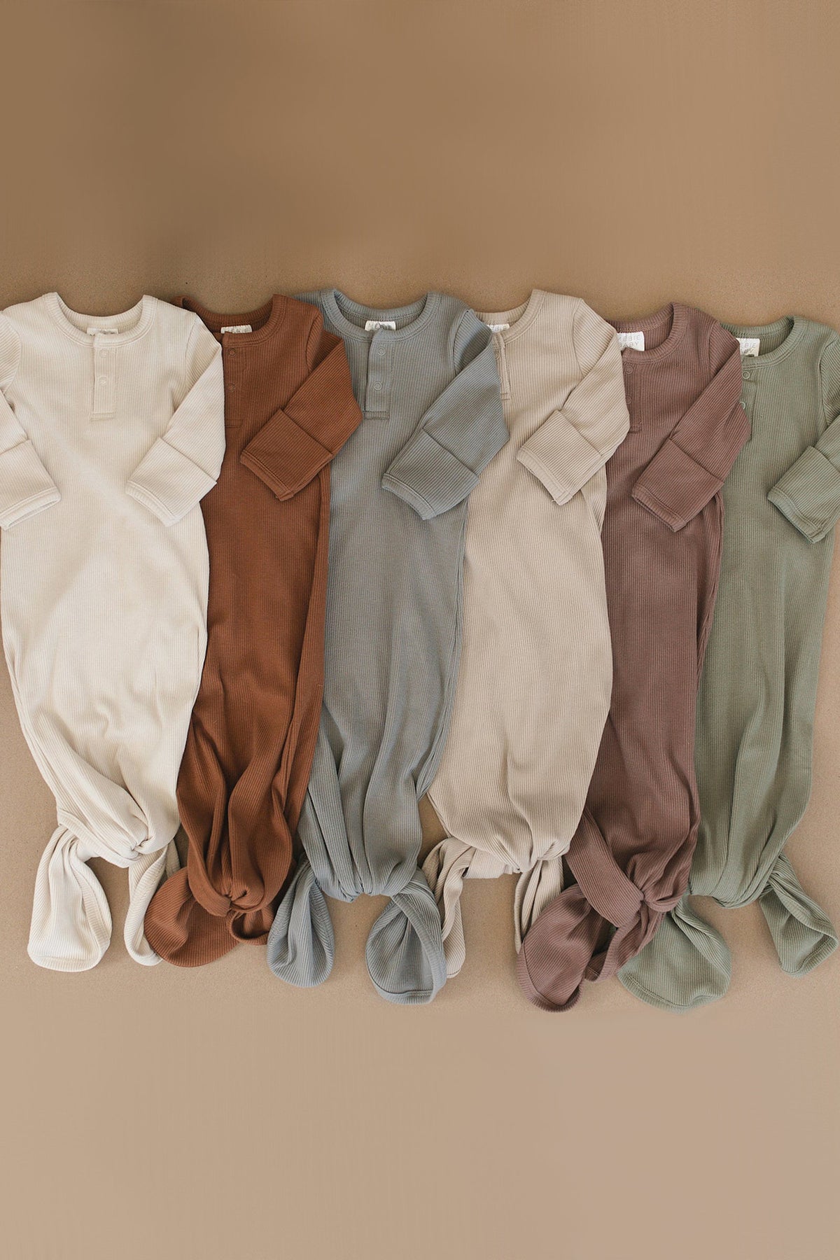Six ribbed baby knot gowns in earthy tones are displayed on a beige background, showcasing organic cotton material.