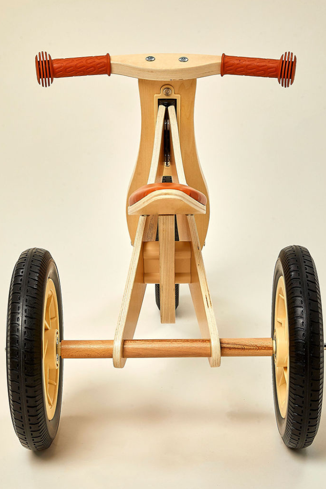 A wooden balance bike with a tricycle design features a natural wood finish, orange rubber grips, and sturdy black tires.