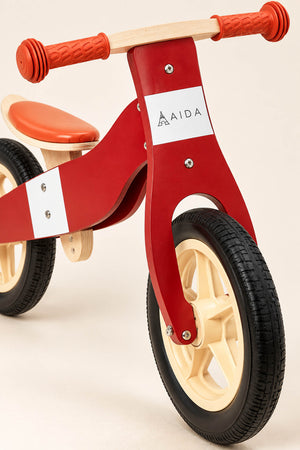A vibrant red wooden balance bike features a sturdy design with an orange seat and cream-colored wheels. Ideal for children aged 3 to 5, it promotes balance and coordination.