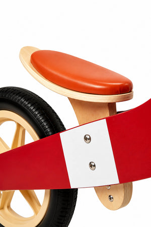 Close-up view of a wooden balance bike featuring a red frame, a natural wood seat, and a brown leather seat cover.