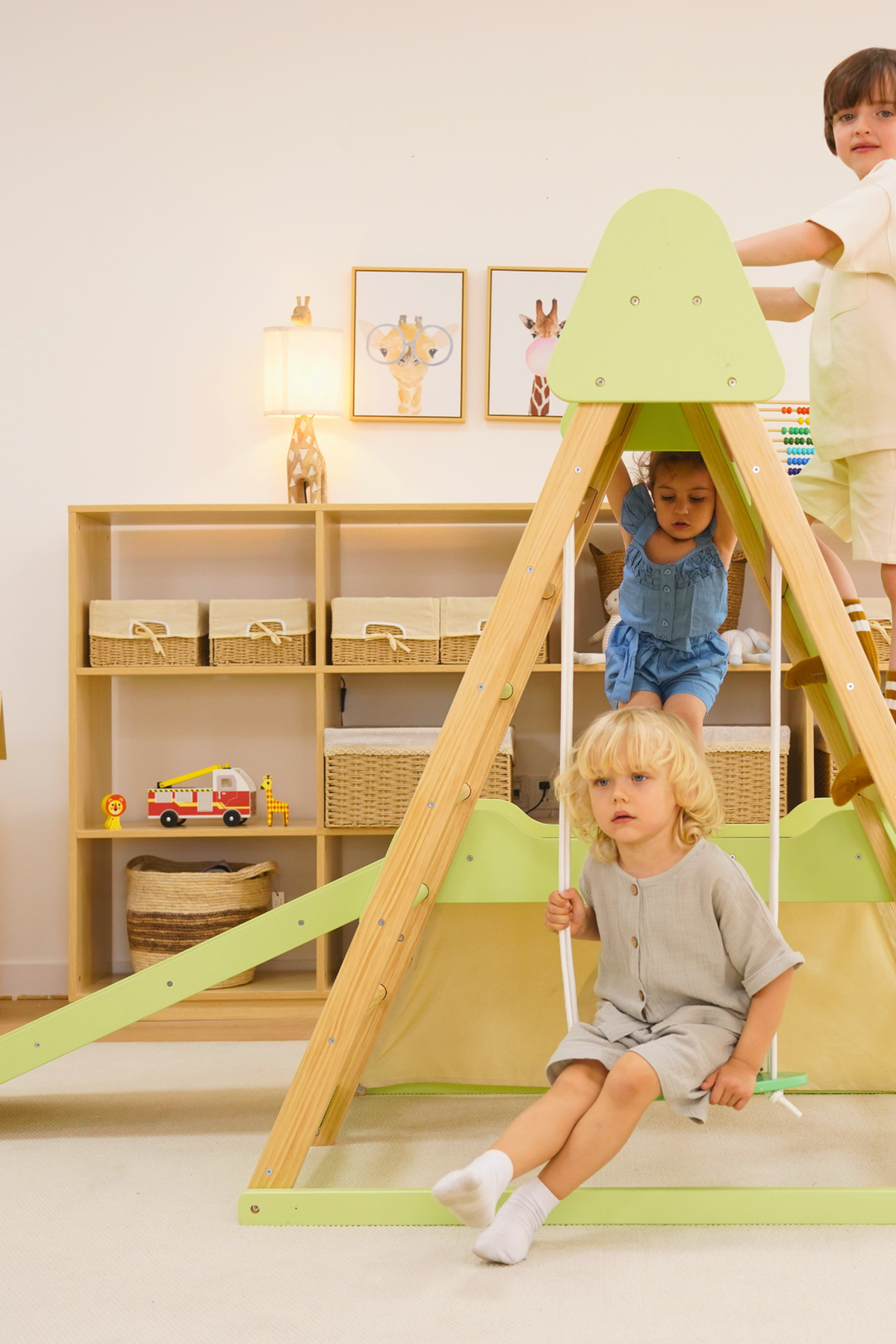Three children play on the Bush 6 in 1 Wooden Jungle Gym Climber, featuring a vibrant green slide and wooden structure. The indoor setting includes stylish storage.