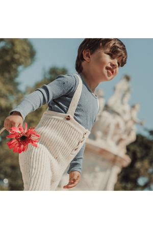 A young child walks outdoors, wearing a light blue ribbed long-sleeve shirt and cream-colored knit overalls with buttons. They hold a pink flower, smiling in a bright, sunny setting.