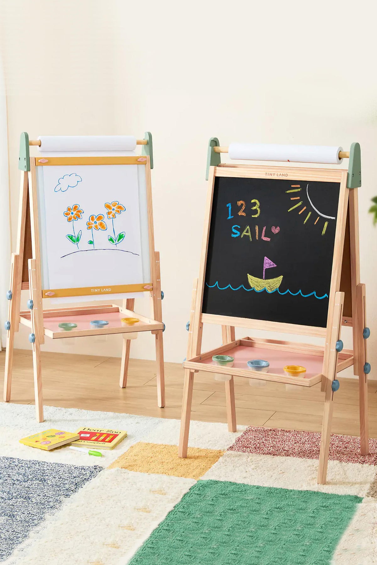 A colorful Tiny Land® 3-in-1 art easel features a magnetic whiteboard on one side and a chalkboard on the other, set in a bright, inviting playroom with a soft rug.